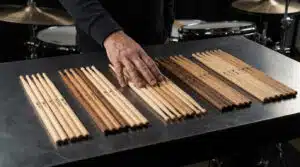 Une main touche des paires de baguettes de batterie variées (bois clair, foncé) alignées sur une table en métal, avec une batterie en arrière-plan.